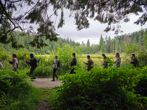 A group of people walk single file along a forest path bordered by dense greenery, under an overcast sky. Tall trees and a grassy wetland stretch out in the background. They all play bamboo buzzers.