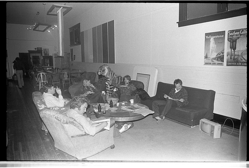 Black-and-white photo of a relaxed lounge area inside a large hall. Several people sit on mismatched couches and chairs arranged around a low coffee table, reading, talking, and drinking. Behind one person in the center is a large tiger sculpture, adding a playful, surreal element. Posters advertising travel destinations hang on the wall behind them, and more tables and chairs extend into the background of the space.