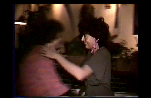 A video still from a VHS recording shows two women hugging in a warmly lit indoor setting. The person on the right is smiling and wearing large 1980s-style earrings. The quality and visual noise (blur and scan lines) are characteristic of analog video formats.