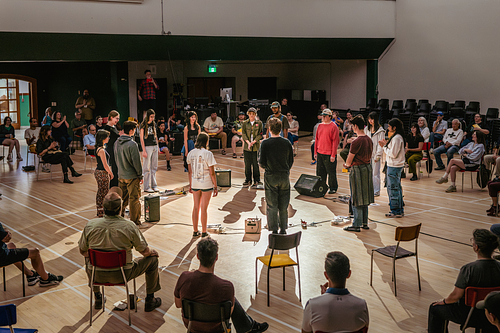 A circle of performers stands among microphones, pedals, and amplifiers in a rehearsal hall, surrounded by seated audience members.