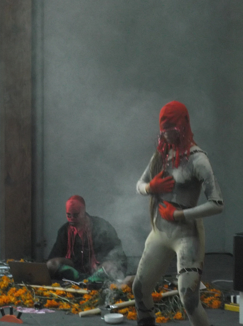A person wearing a red balaclava, red gloves, and a distressed grey bodysuit stands with their hands resting over their chest and stomach. Behind them, another person in a similar red face covering sits cross legged on the floor in front of an open laptop. Bunches of orange flowers and other objects are strewn on the floor. 
