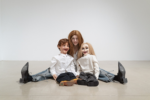 A person with a long brown wig, white collared shirt, blue jeans, and black pointy toed shoes sits on a gallery floor with their legs spread wide, holding two ventriloquist puppets. One has short tousled brown hair and the other has long blonde hair. They are both dressed in white shirts. 
