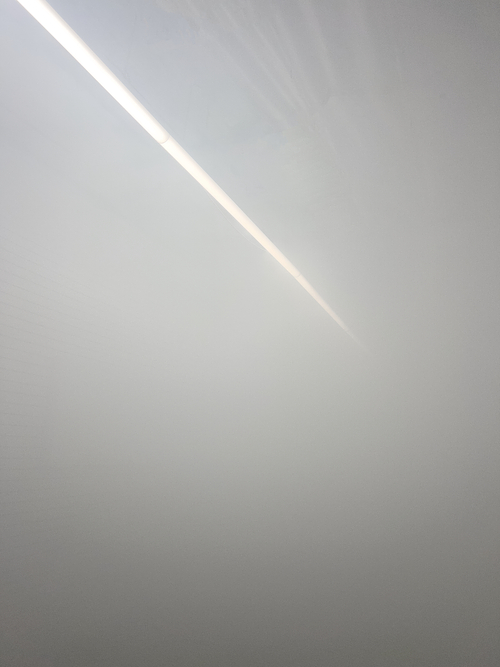 A vertical photograph of a brightly lit, fog-filled interior space where thick white haze obscures most details, with only a single diagonal strip of ceiling light visible from the top left toward the center right against an otherwise smooth, pale gray background.