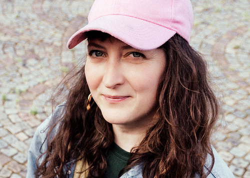 A portrait of Eve Risser wearing a pink baseball cap and gold hoop earrings, smiling slightly. She is outdoors on a cobblestone path, dressed casually in a denim jacket.