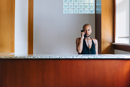 Chipo Chipaziwa stands behind a reception desk holding a black telephone to her ear. She wears a black halter-top with a deep neckline and meets the camera's gaze directly. The desk has a speckled marble top, and the background features light wooden panels and a glass block window, allowing soft daylight to filter into the space. 