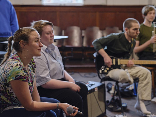 Four young adults sit in a semi-circle in a wood-paneled room, participating in a music session. In the foreground, a person in a colorful shirt holds a microphone and smiles attentively. Beside them, another person sits quietly, while a third plays an electric guitar, and a fourth holds a brass instrument. Music equipment is visible on the floor. The atmosphere appears focused and collaborative.