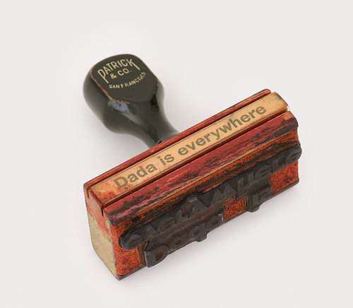 An image of a wooden rubber stamp with the phrase "Dada is Everywhere" printed on it.