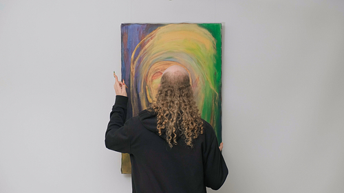 Archie Barry wears a black hoodie and stands facing a colorful painting with swirls of yellow, green, and purple. They hold up one hand with fingers slightly raised, appearing to engage with the artwork.