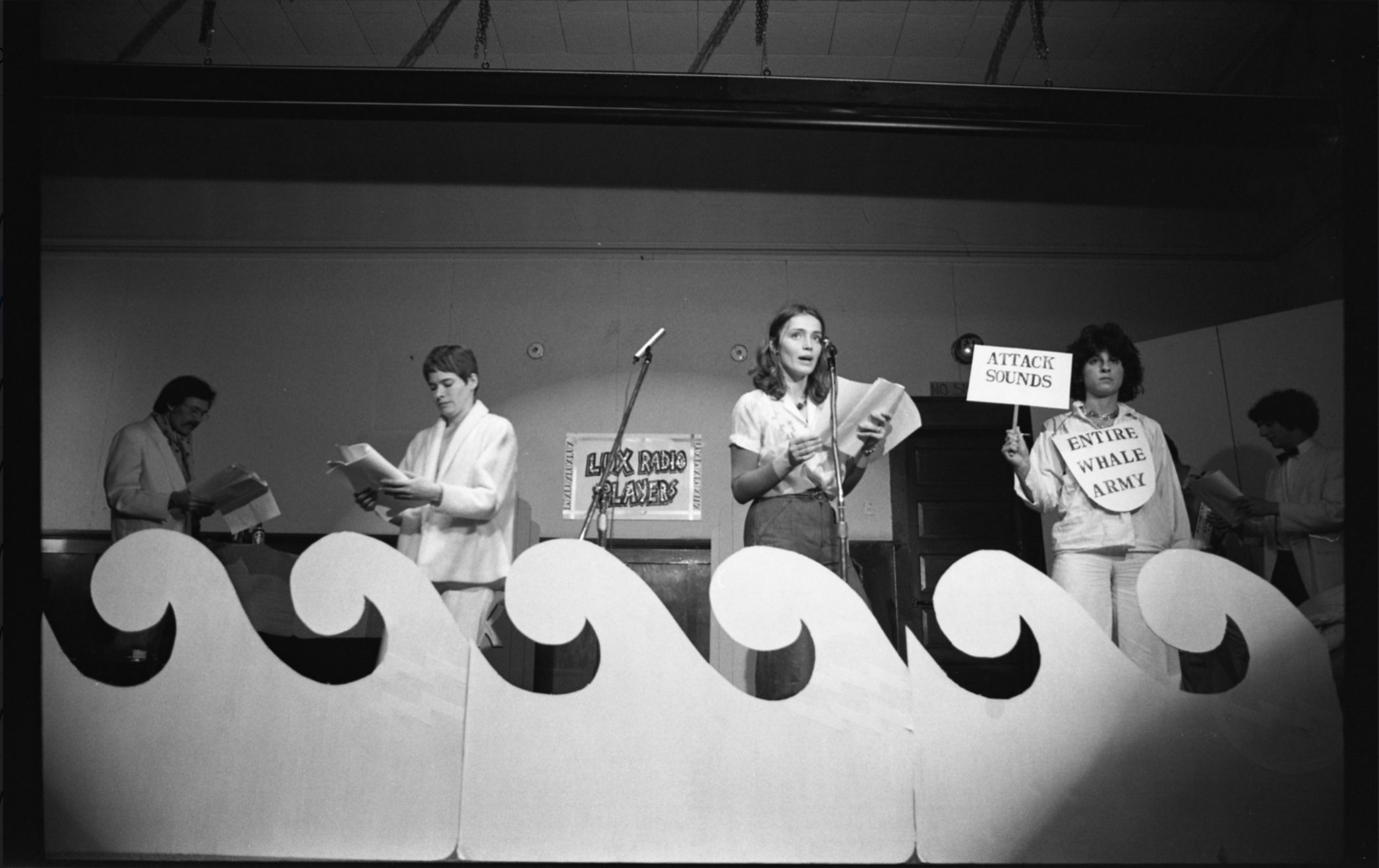A black and white photograph in which four performers stand in a row behind cardboard cut-outs of ocean waves. Robert Amussen and Kate Craig look down at scripts, while Suzanne Ksinan stands in a spotlight and recites. Martha Miller looks holds a sign that reads “Attack Sounds” and wears another sign on her chest that reads “Entire Whale Army.”