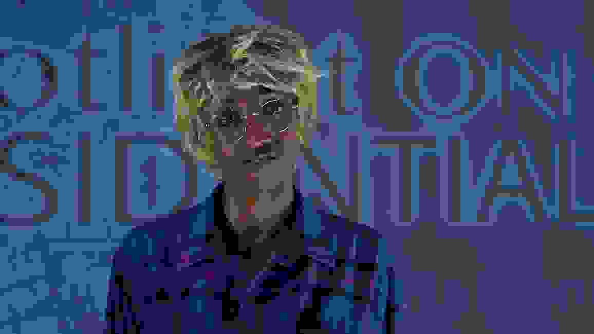 Lex Brown, wearing a blue plaid shirt, glasses, and cropped blonde wig, stands in front of a blue background of layered digital images including a satellite photograph of buildings near a river.